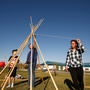 Three people are assembling a teepee frame with wooden poles and string in an outdoor setting under a clear blue sky.