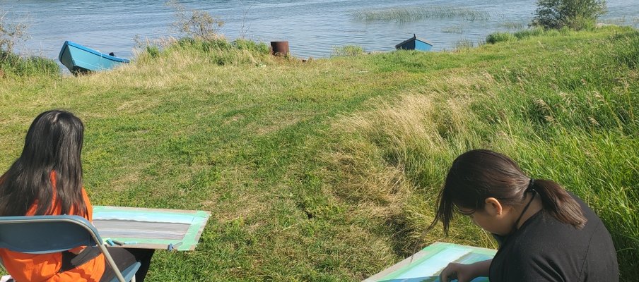 Two young people from English River First Nation’s sitting on chairs in the grass, in front of water, taking part in Summer Land-Based Learning.