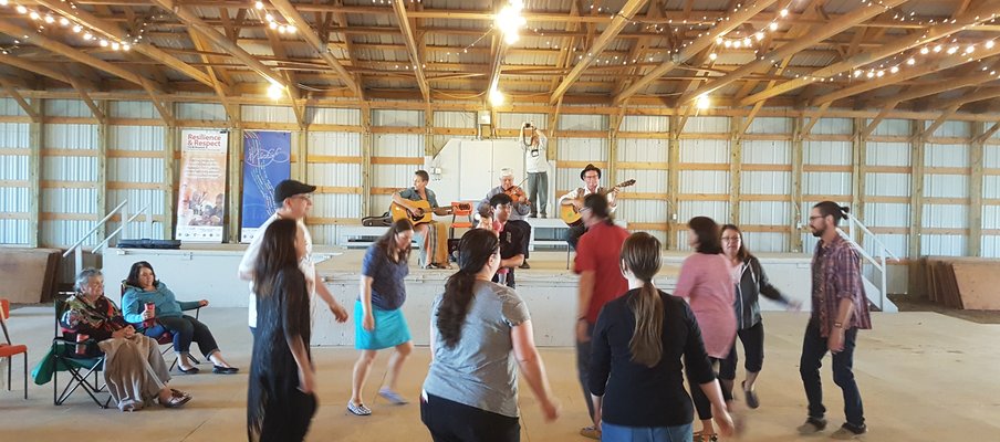 A small crowd at the John Arcand Fiddle Festival, gathered in front of a stage where three musicians are playing.