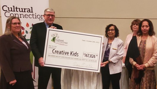 Three people posing in front of a Cultural Connections banner. Two of them are holding a big cheque with the Cultural Connections logo, which reads "Pay to the order of Creative Kids," for the amount of $167,826.