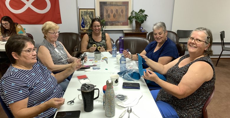 Five people sit around a table engaged in a crafting activity. Various crafting materials, including scissors and thread spools, are spread on the table. A red flag with a white infinity symbol is on the wall.