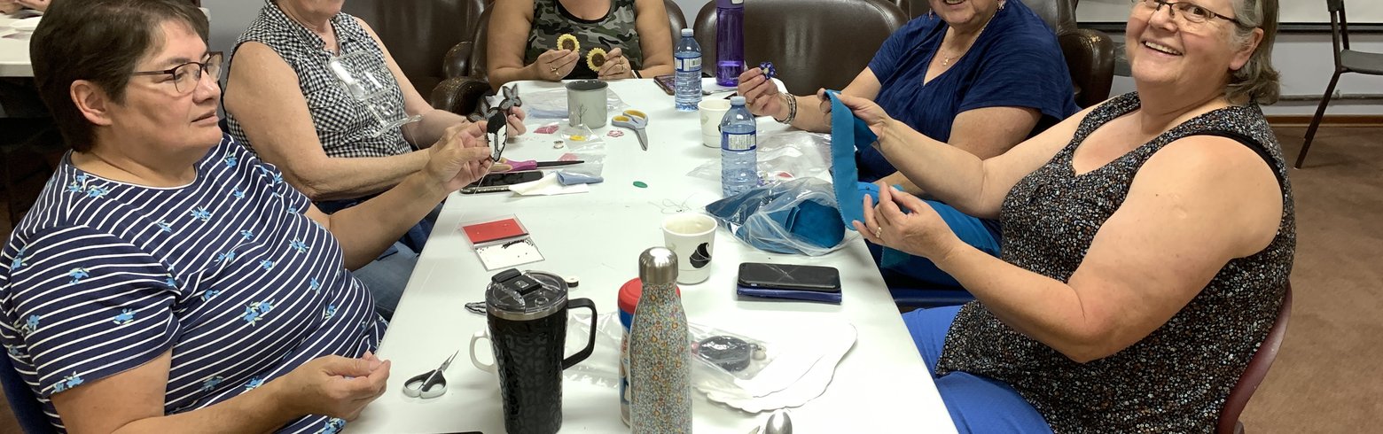 Five people sit around a table engaged in a crafting activity. Various crafting materials, including scissors and thread spools, are spread on the table. A red flag with a white infinity symbol is on the wall.