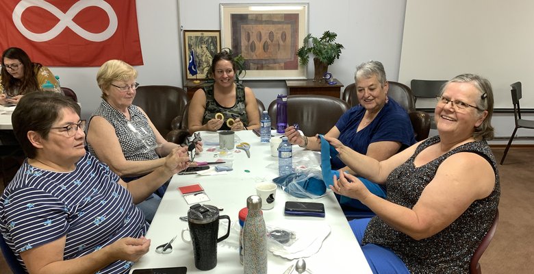 Five people sit around a table engaged in a crafting activity. Various crafting materials, including scissors and thread spools, are spread on the table. A red flag with a white infinity symbol is on the wall.