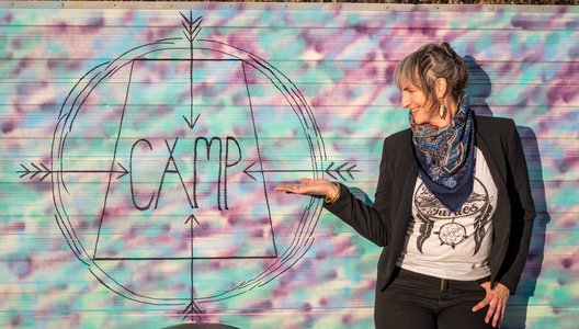 Eliza Doyle, volunteer artistic director of CAMP Sask Arts, standing pointing to a colourful sign that reads: "CAMP."