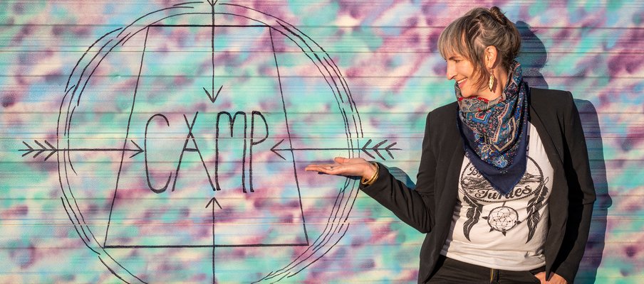 Eliza Doyle, volunteer artistic director of CAMP Sask Arts, standing pointing to a colourful sign that reads: "CAMP."