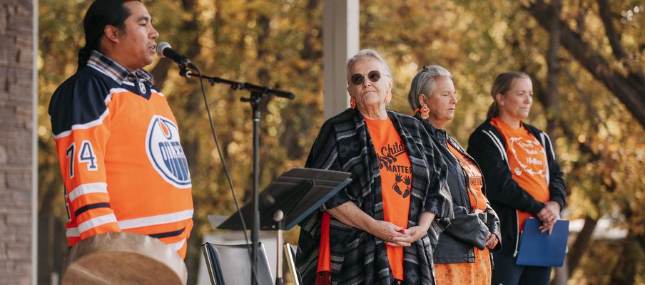 A group of four people wearing orange clothing. On the left, Lindsay Littlechief talks on a mic about the significance of the Honour song. Elder Angie McArthur-Delorme is second from the right.