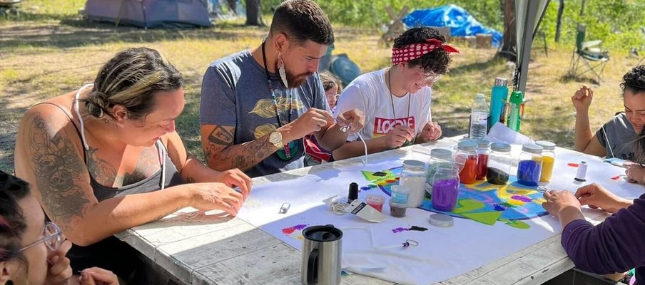 Participants and Knowledge Keepers from this year’s OUT on the Land camp sitting around table beading. The background shows trees and camping tents.