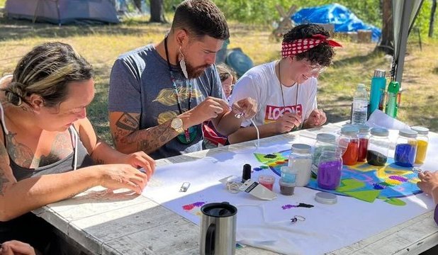 Participants and Knowledge Keepers from this year’s OUT on the Land camp sitting around table beading. The background shows trees and camping tents.