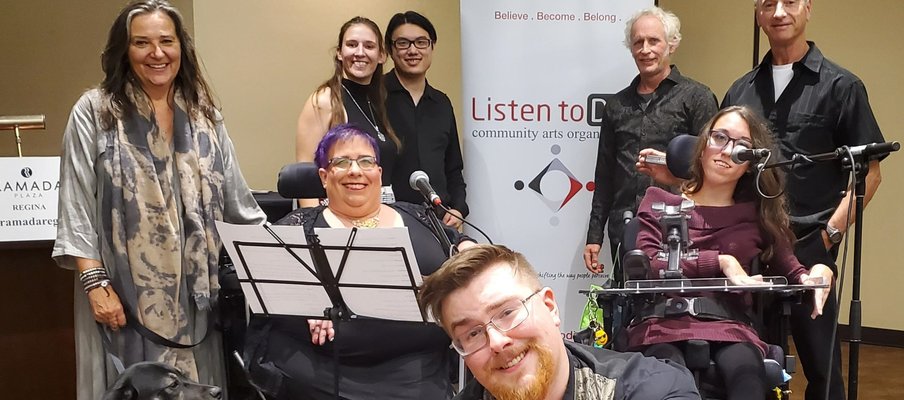 Eight people posing in front of a Listen to Dis' banner, smiling at the camera, also joined by a service dog in training. Two of the people in the photo are seated in wheel chairs and have microphones; one person in squatting; the rest are standing.