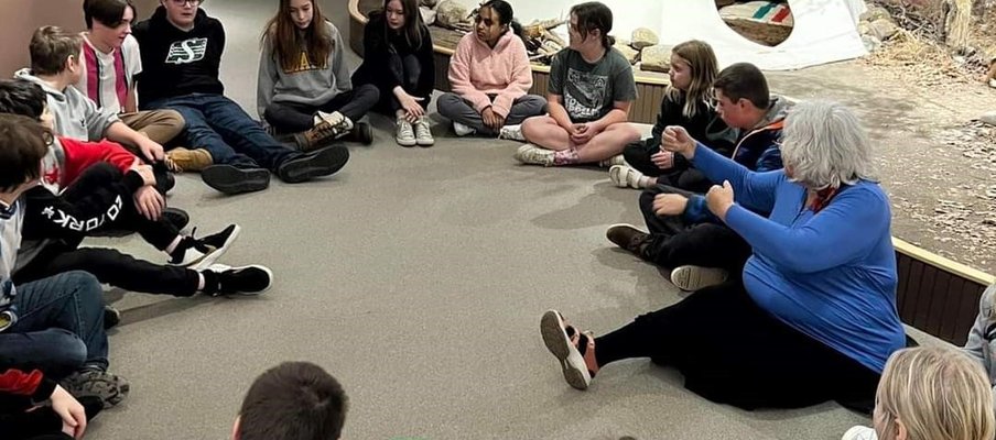 A photo of, Barb Frazer, Knowledge Holder, and a group of kids, all sitting on the floor in a circle. In the background there is a tipi.