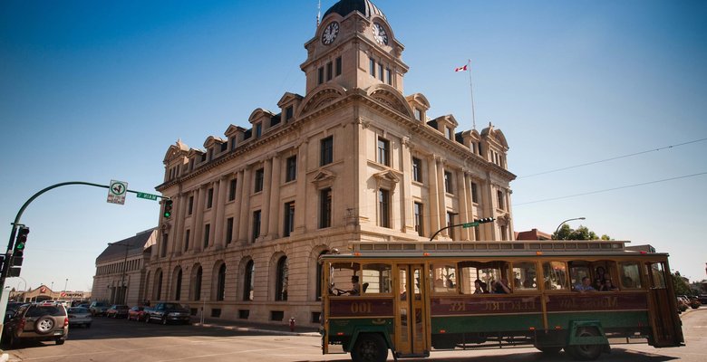 A photo of a trolley driving in front of a building in Moose Jaw.
