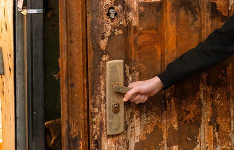 a hand turns a door handle and opens a damaged brown door