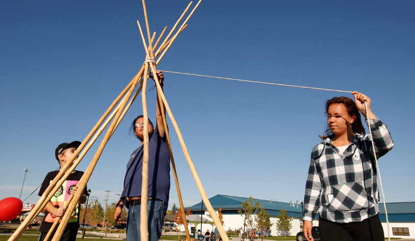 Three people are assembling a teepee frame with wooden poles and string in an outdoor setting under a clear blue sky.