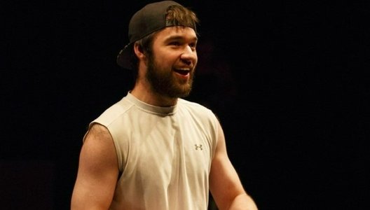 A photo of John Loeppky performing in a play. Loeppky is barefoot and wearing athletic wear, sitting on a wheelchair.