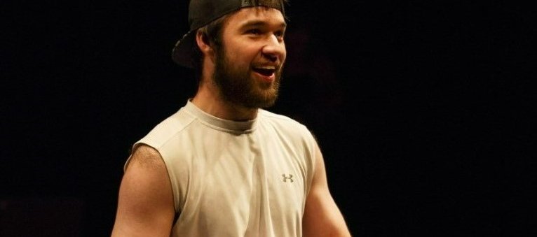 A photo of John Loeppky performing in a play. Loeppky is barefoot and wearing athletic wear, sitting on a wheelchair.