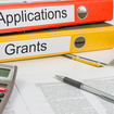 Two binders labeled "Applications" and "Grants", a calculator, and a pen on top of documents.