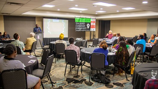 A photo of PRIMORG's first event. it shows people gathered in an events room, looking at a speaker at the podium. Next to the speaker there is a poster that reads: The Afro-Canadian Community.