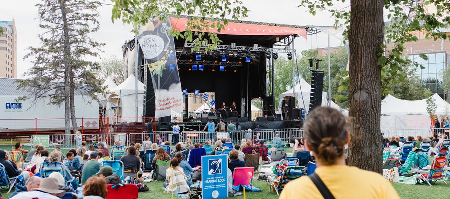 A photo of the crowd gathered on the lawn in front of a big festival stage. A light blue sign in the middle of the lawn advertises a hearing loop.