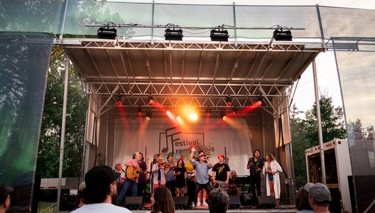 A photo of a group of performers on a festival mainstage. The performers are holding instruments and singing into microphones. In the back there is a big sign that reads "Festival Fransaskois."