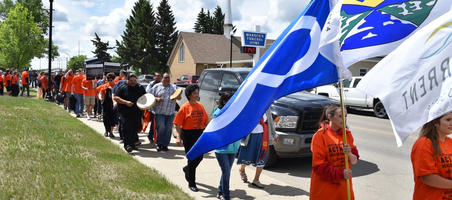 A crowd of people wearing orange shirts, marching with posters, drums and flags, including the Métis flag.