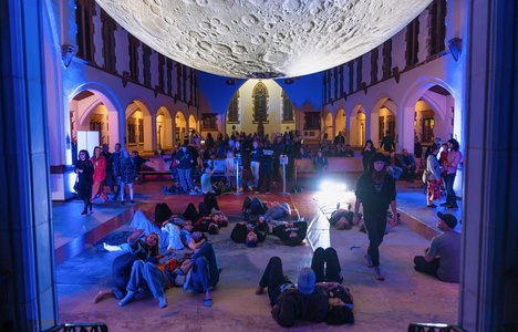 A photo of people sitting down looking at a big art installation of a moon at Nuit Blanche YXE 2023.