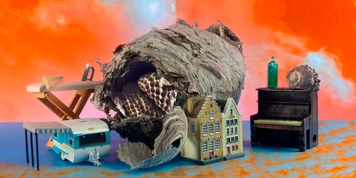 A collection of objects is arranged over a dreamy orange and blue background. Playing with scale, a wasp nest is laid over miniature rowhouses, ironing board, camper trailer, a piano, a shell, and a bottle.